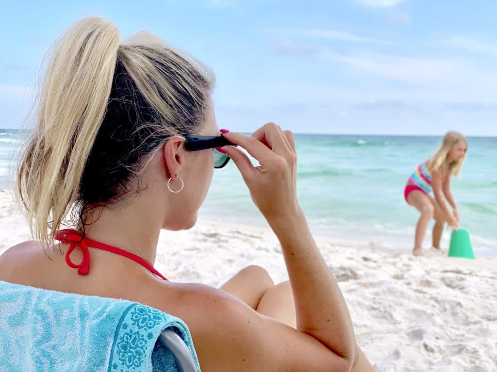 Woman wearing dark sunglasses at the beach while her daughter plays in the sand.
