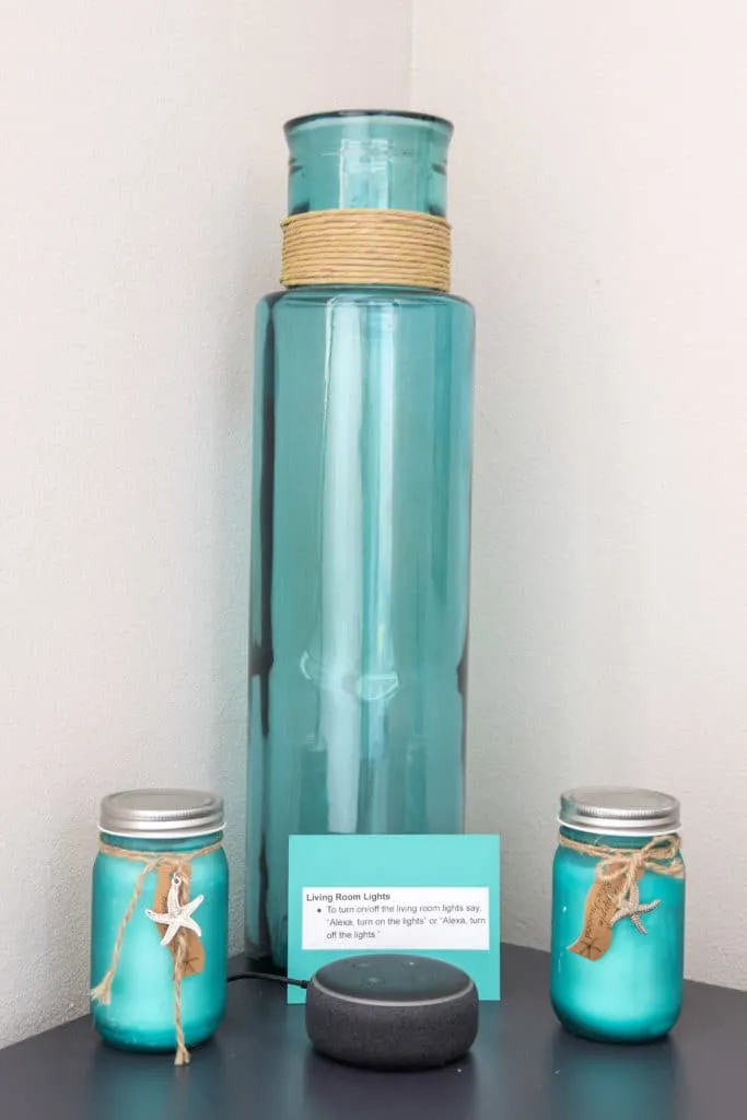 Teal glass jars, candles, and Alex Echo Dot on shelf in living room with sign on how to use Alexa Echo Dot to control lights.
