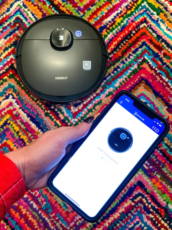 Woman's hand holding phone with robotic vacuum app in front of colorful rug with robotic vacuum.