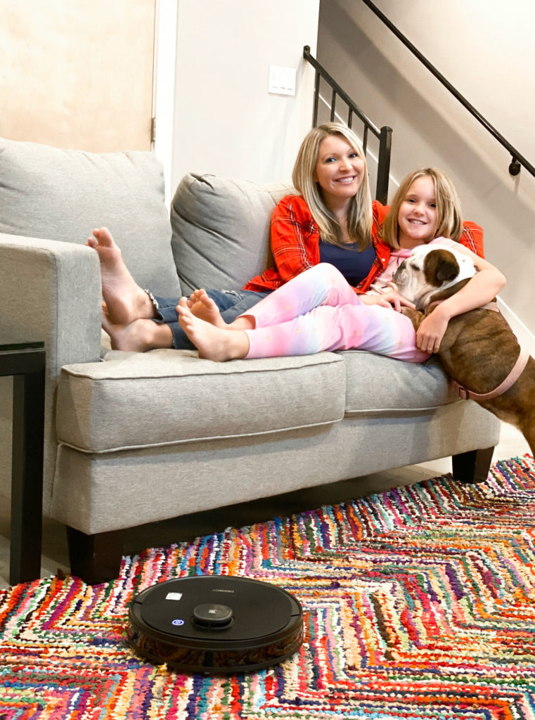 Mom and daughter on couch with dog on colorful rug and robotic vacuum.